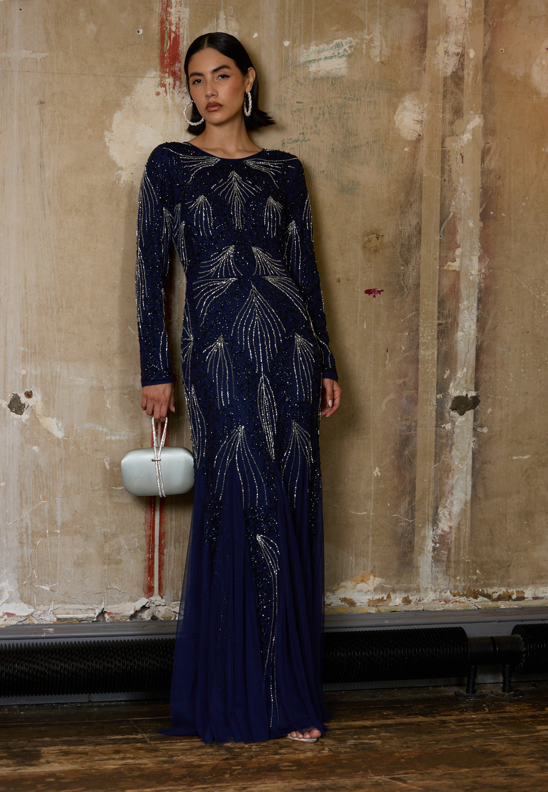Woman in a long, dark blue evening gown with intricate patterns against a textured wall.