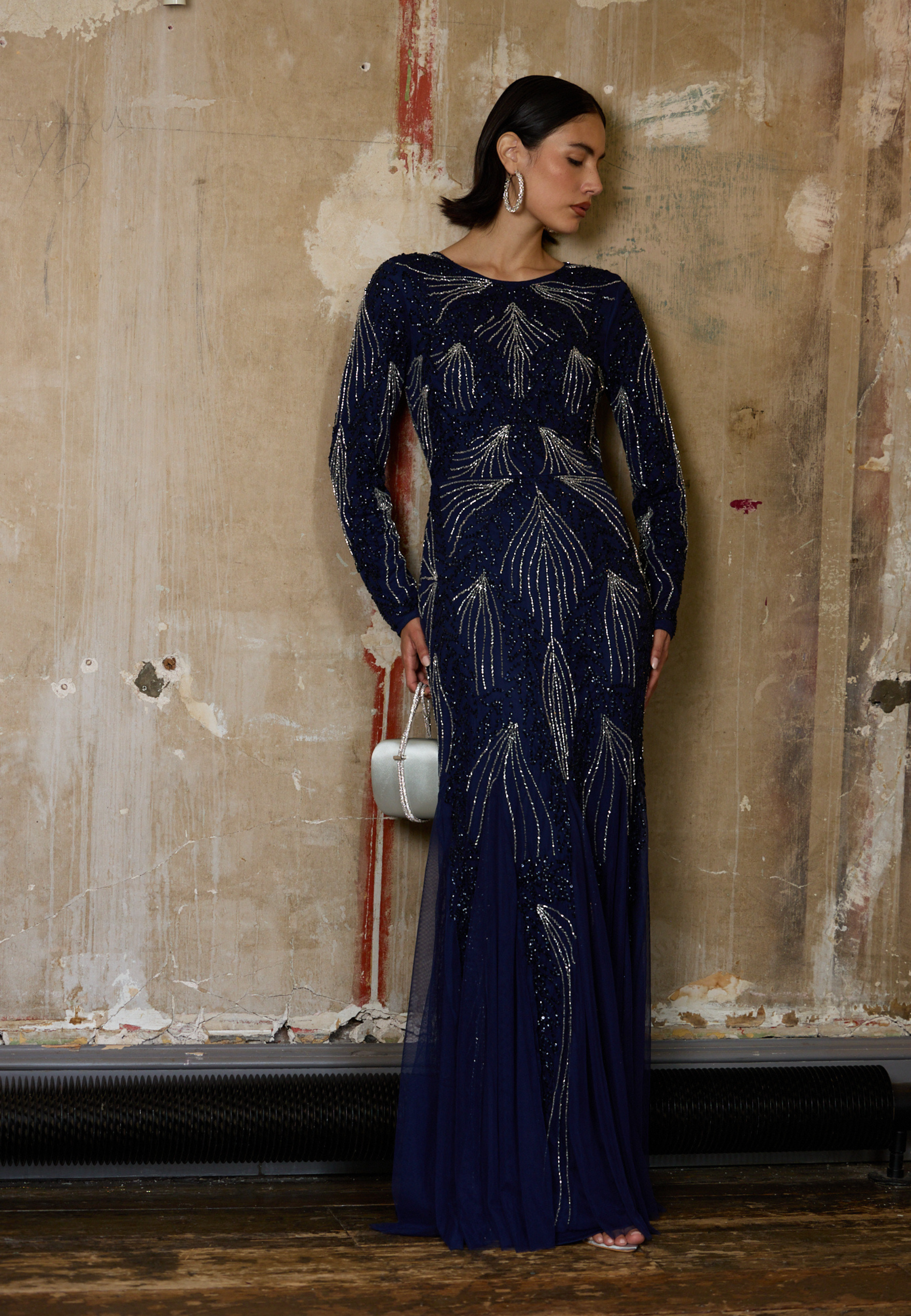 Woman in a long, dark blue evening gown with feather patterns against a textured wall.