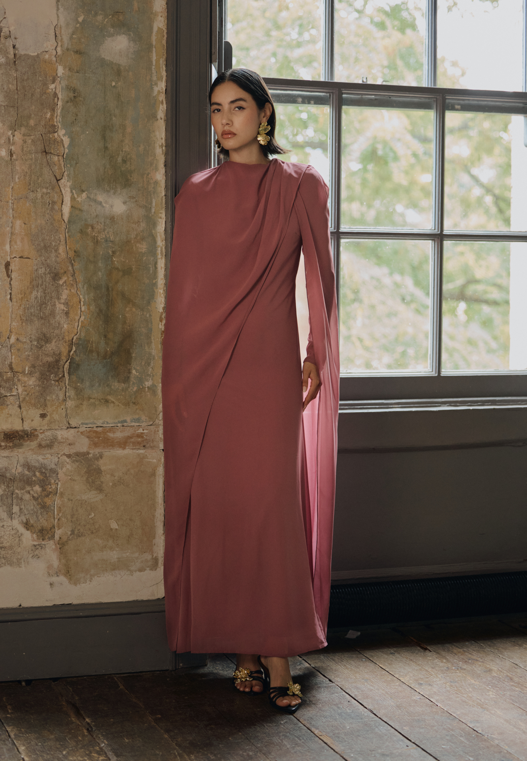Woman in a long, flowing dress standing in a room with a window and textured wall.