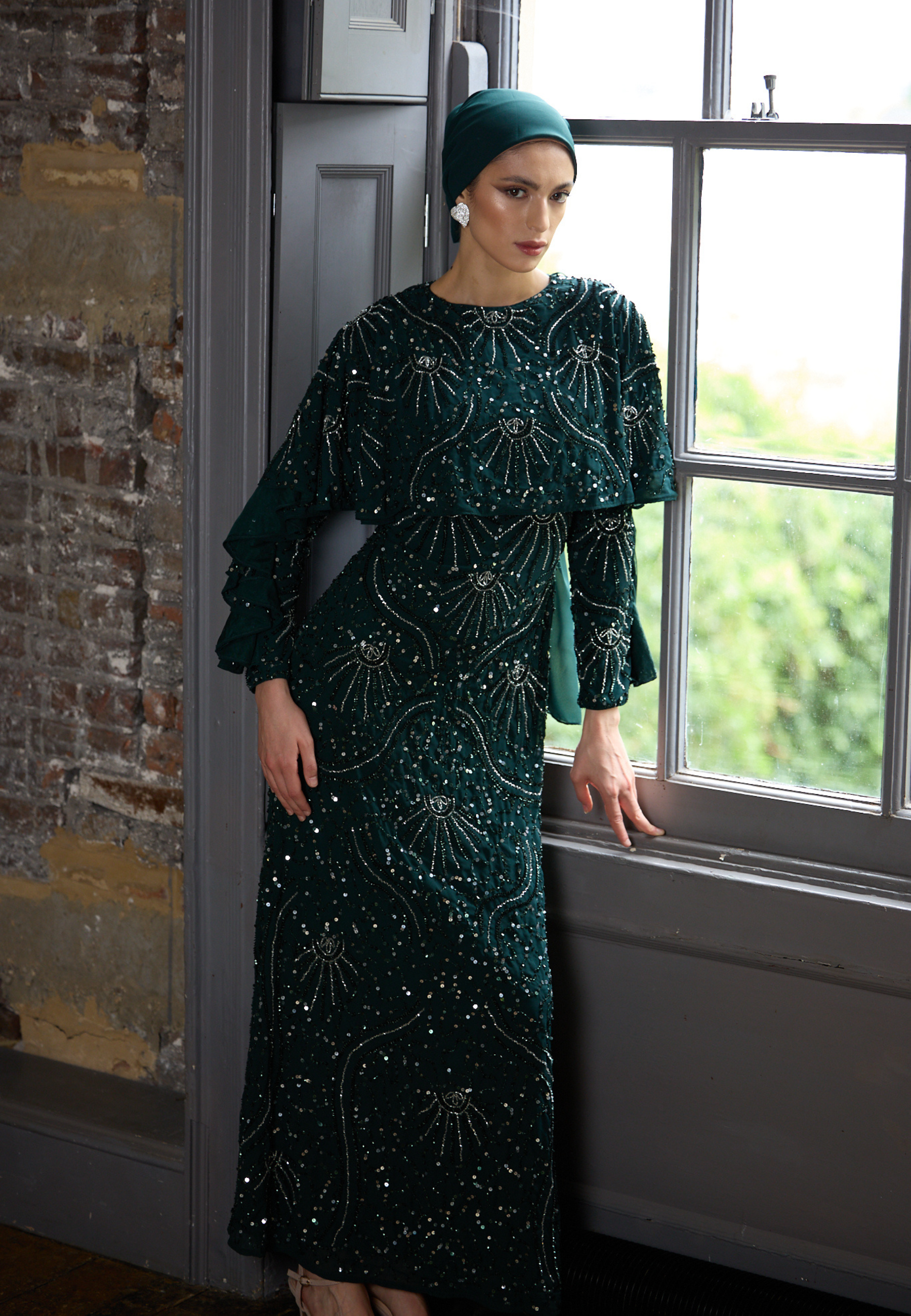 Woman in a green dress with intricate patterns standing by a window.
