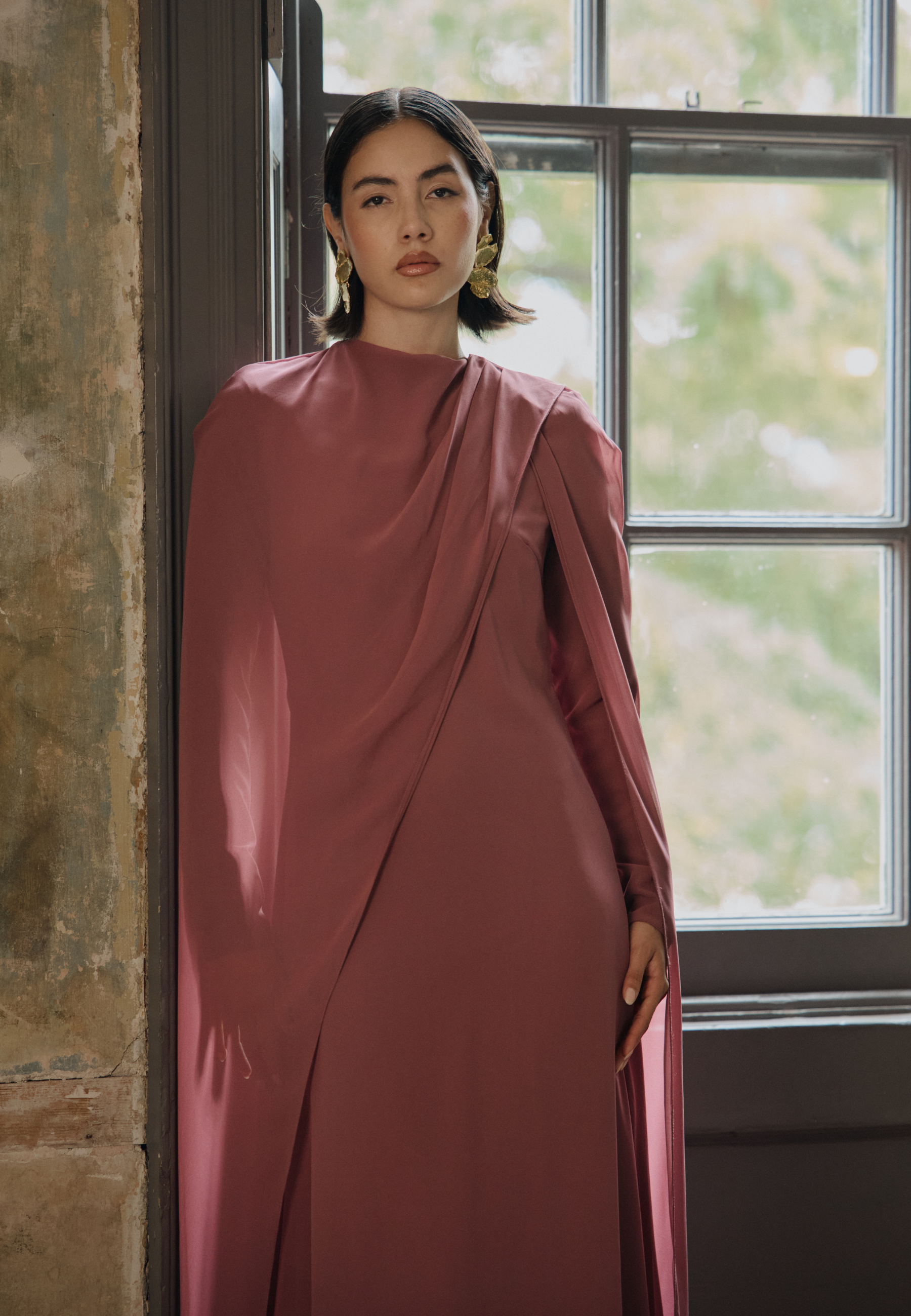 Woman in a mauve dress standing by a window with a blurred outdoor background