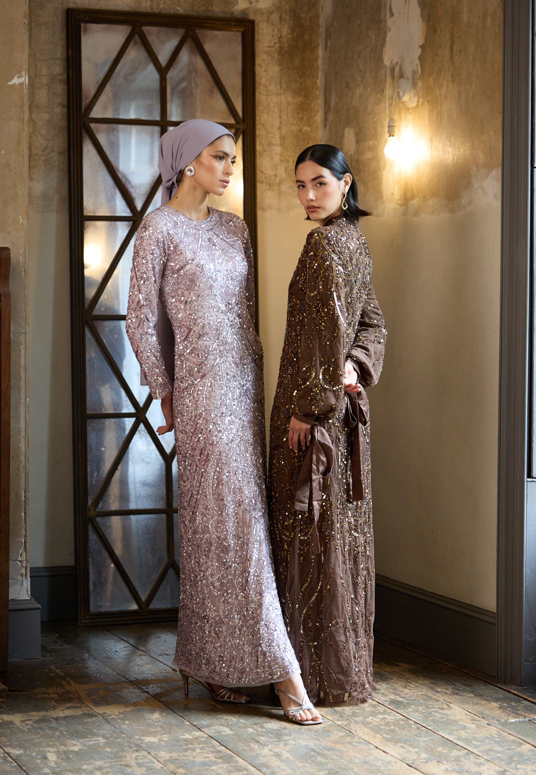 Two women in elegant dresses standing in a room with a decorative mirror.