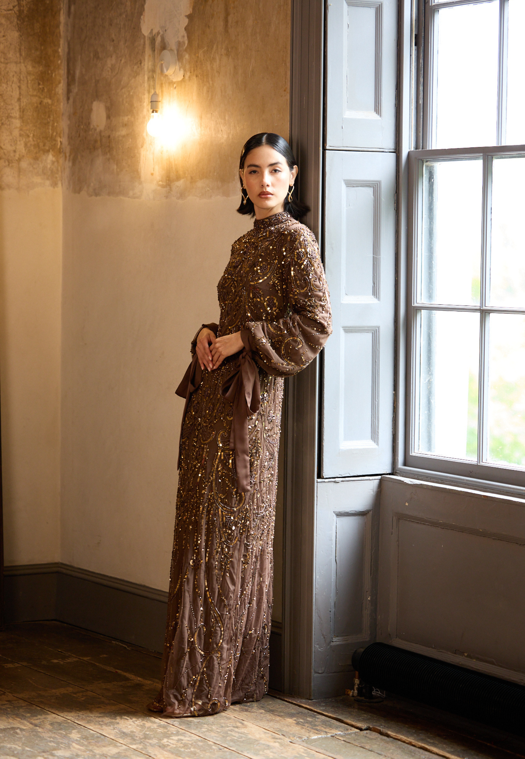Woman in a long, brown sequined dress standing in a room with a window and light fixture.
