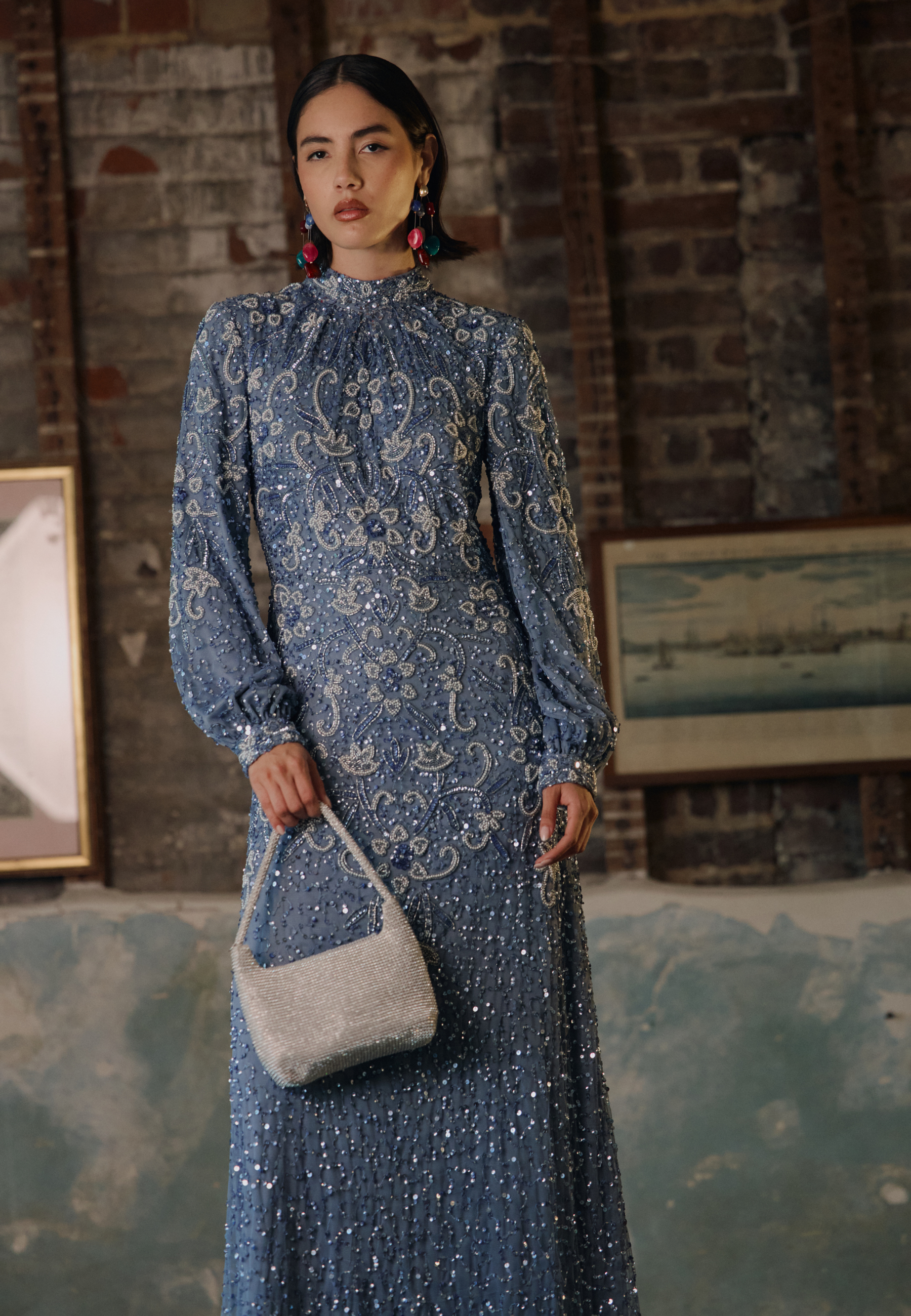 Woman in a blue patterned dress holding a gray handbag against a brick wall background.