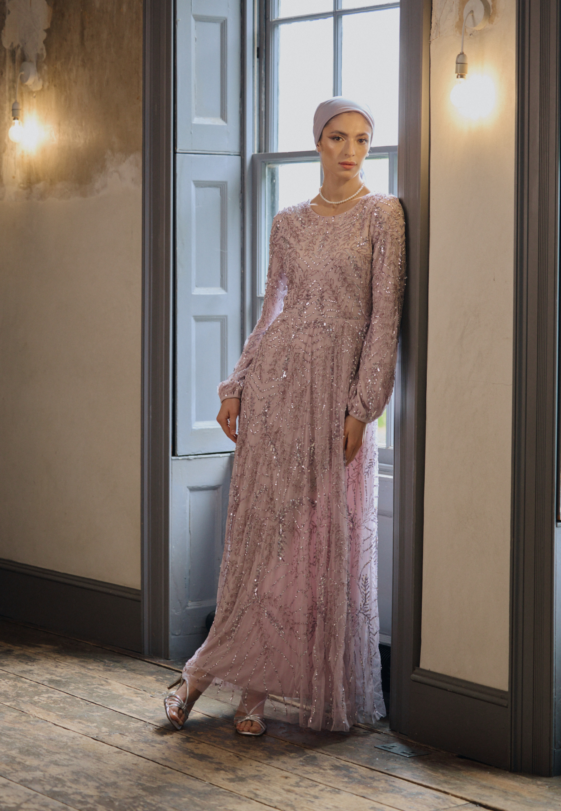 Woman in a long, light pink dress standing in a room with wooden floor and white walls.