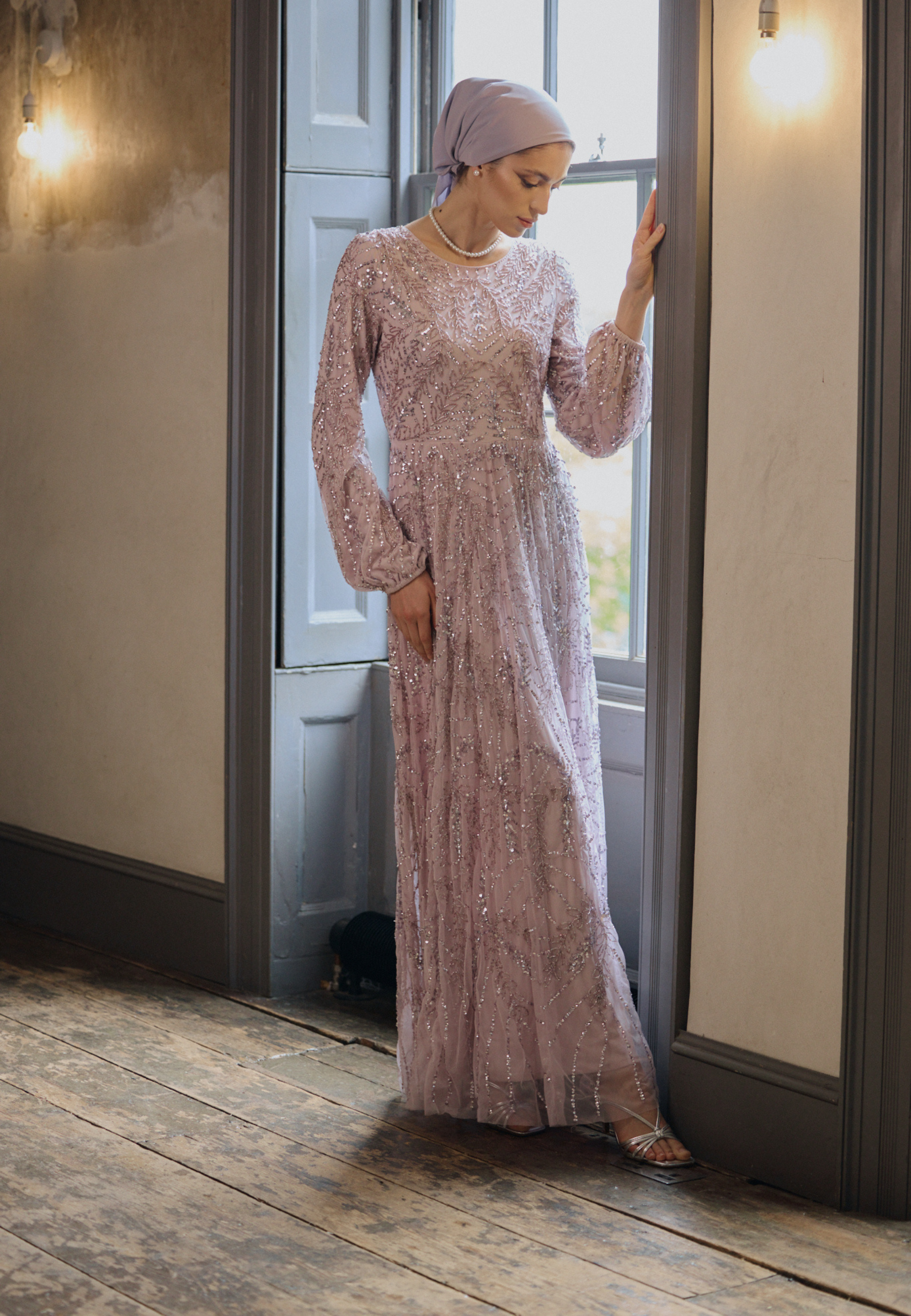 Woman in a long, light pink lace dress standing in a room with wooden floor and large window.
