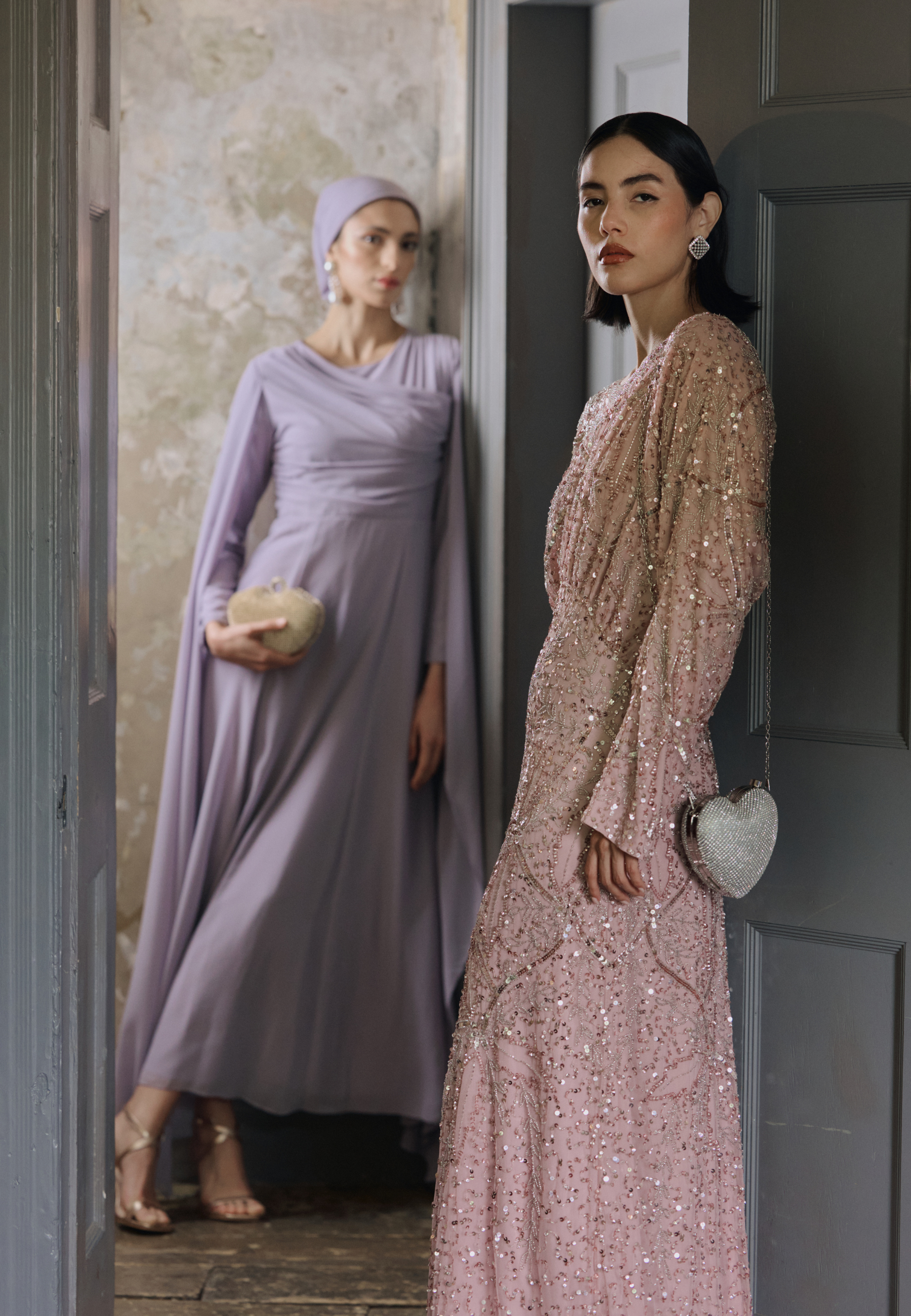 Two women in elegant dresses standing in a doorway, one in lavender and the other in pink.