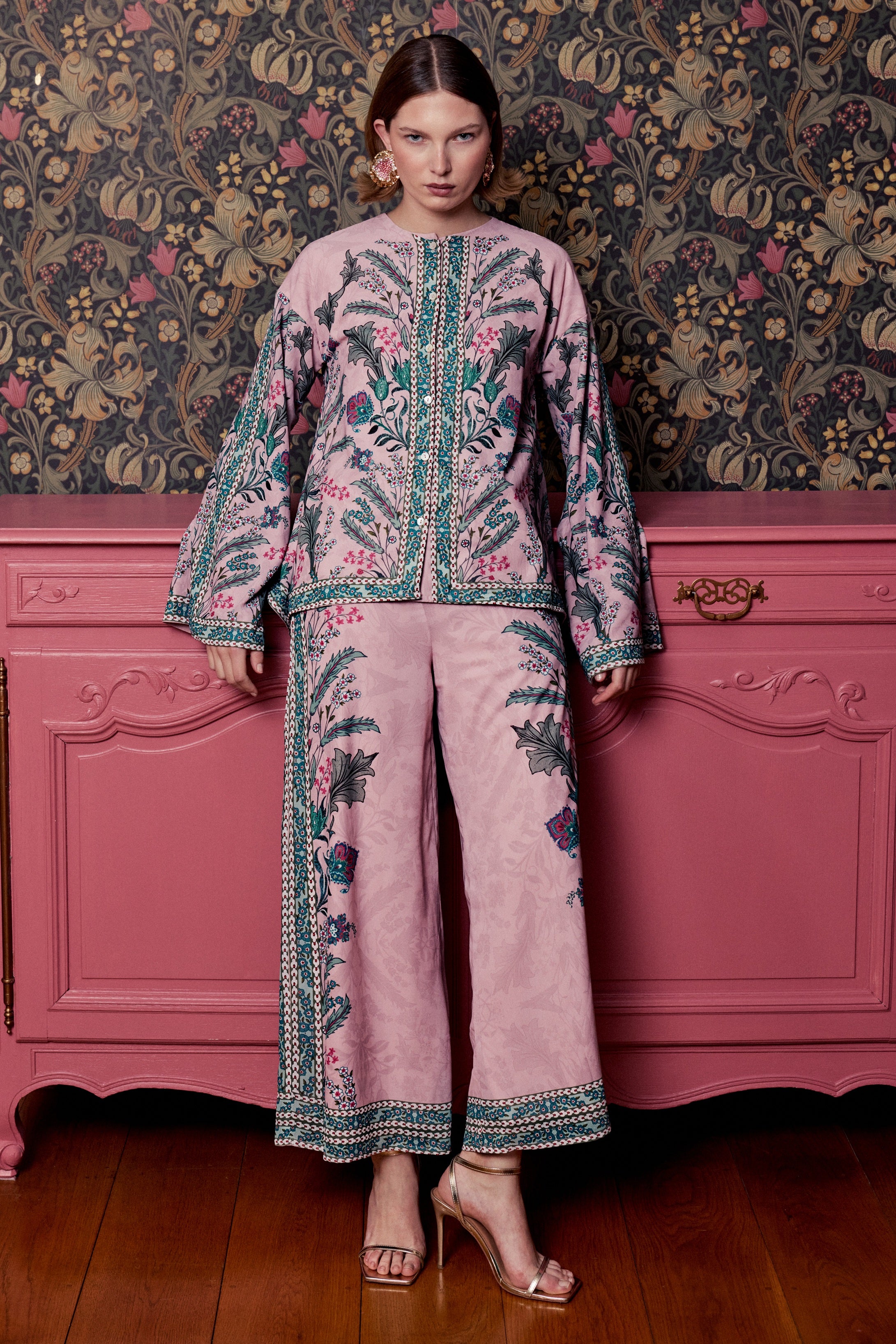 Woman wearing a floral embroidered outfit standing in front of a pink dresser with decorative wallpaper.