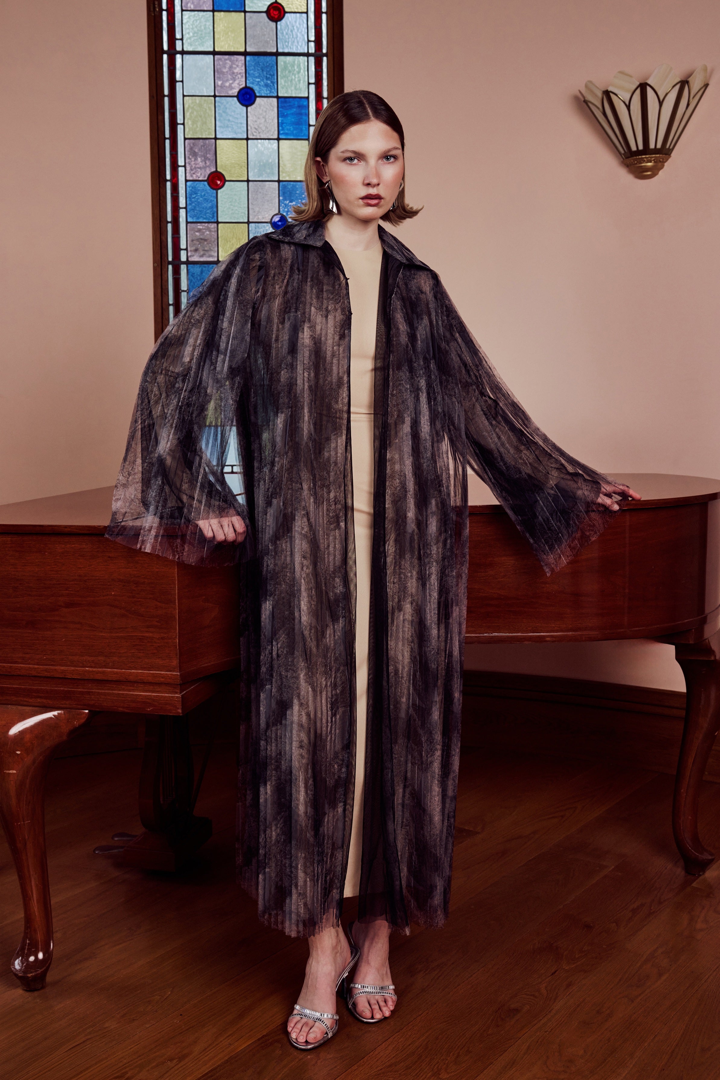 Woman in a long, dark coat standing in a room with a stained glass window and wooden furniture.