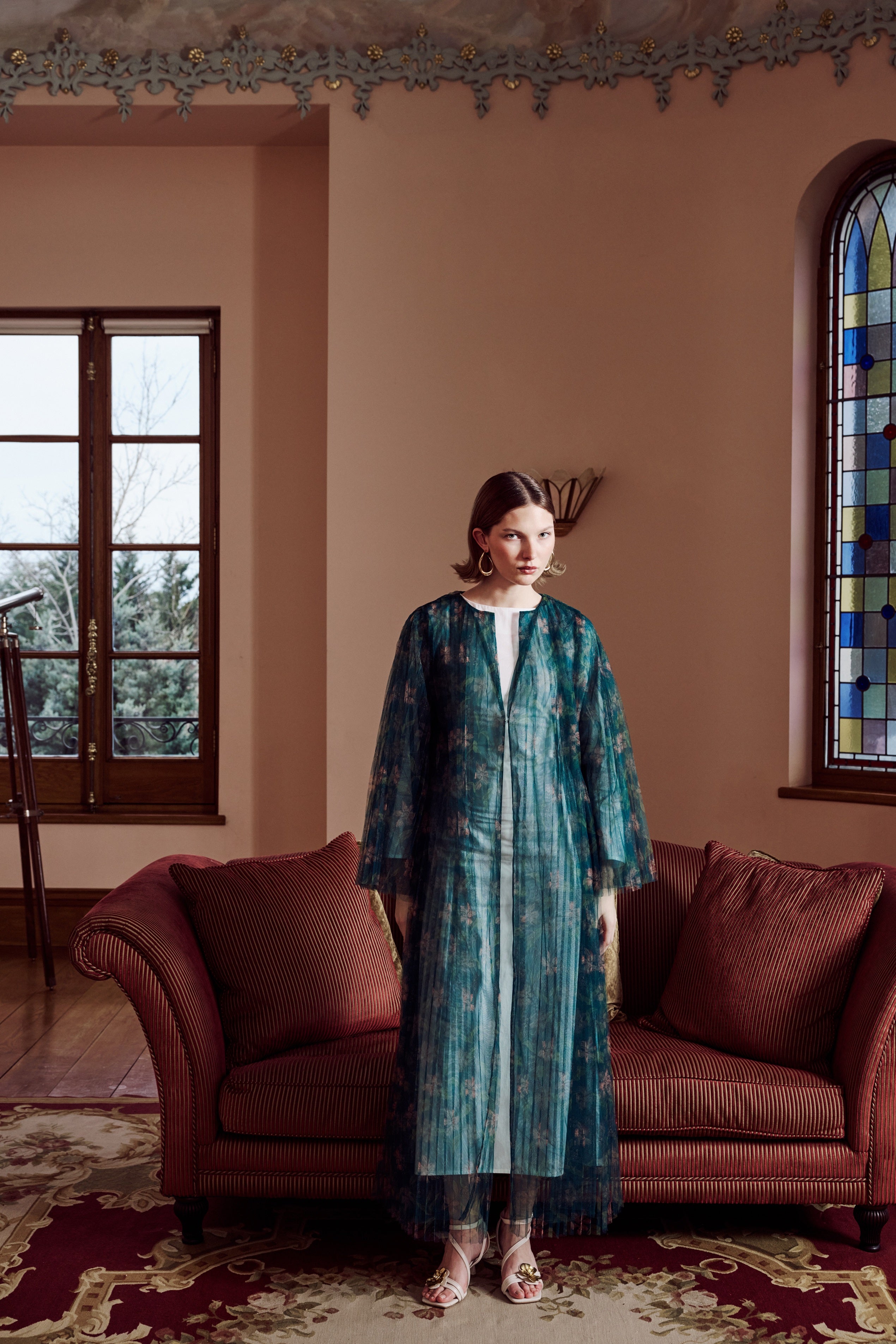 Woman in a teal dress standing in a room with a brown couch and stained glass window.