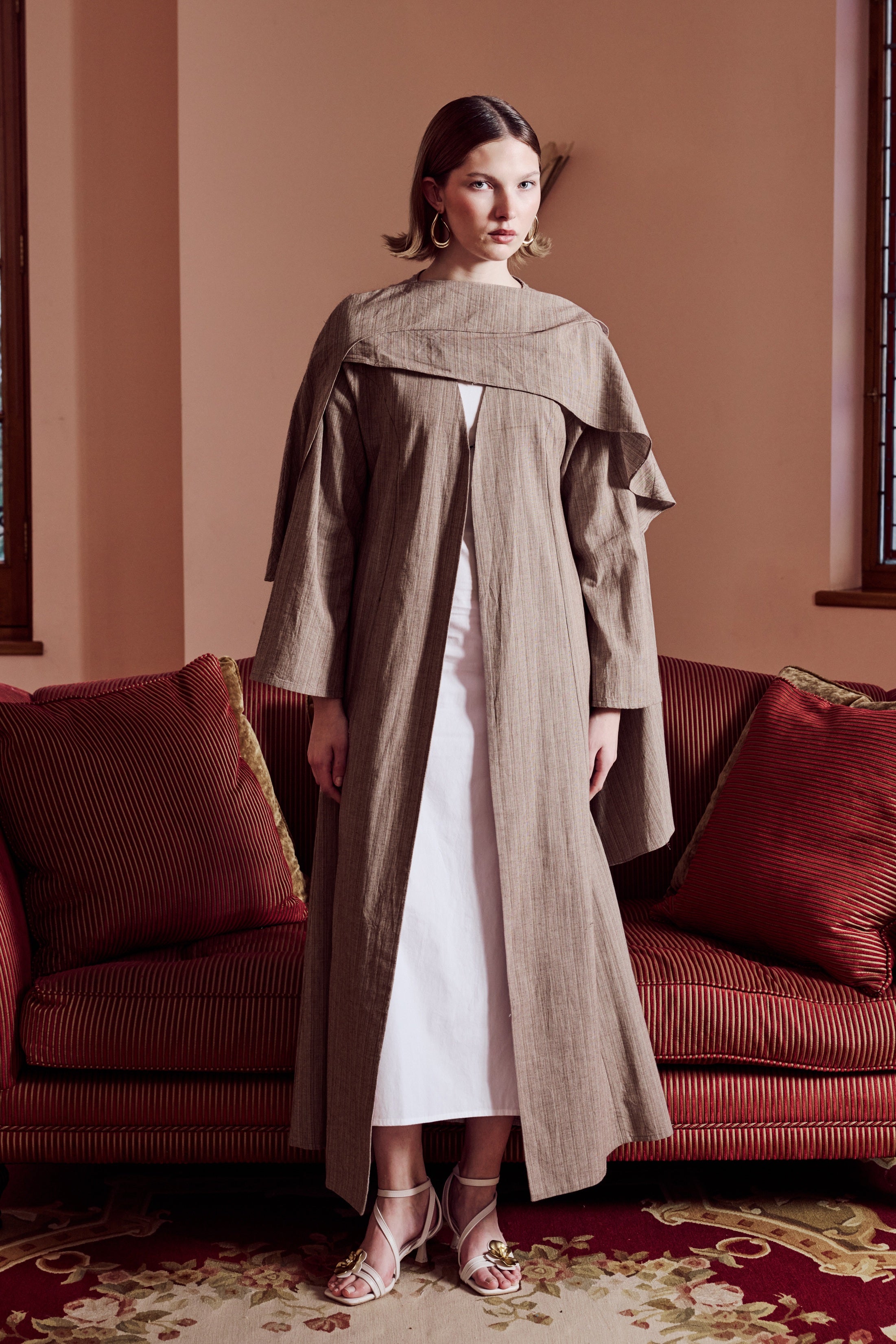 Woman wearing a long beige coat over a white dress in a room with a red sofa.