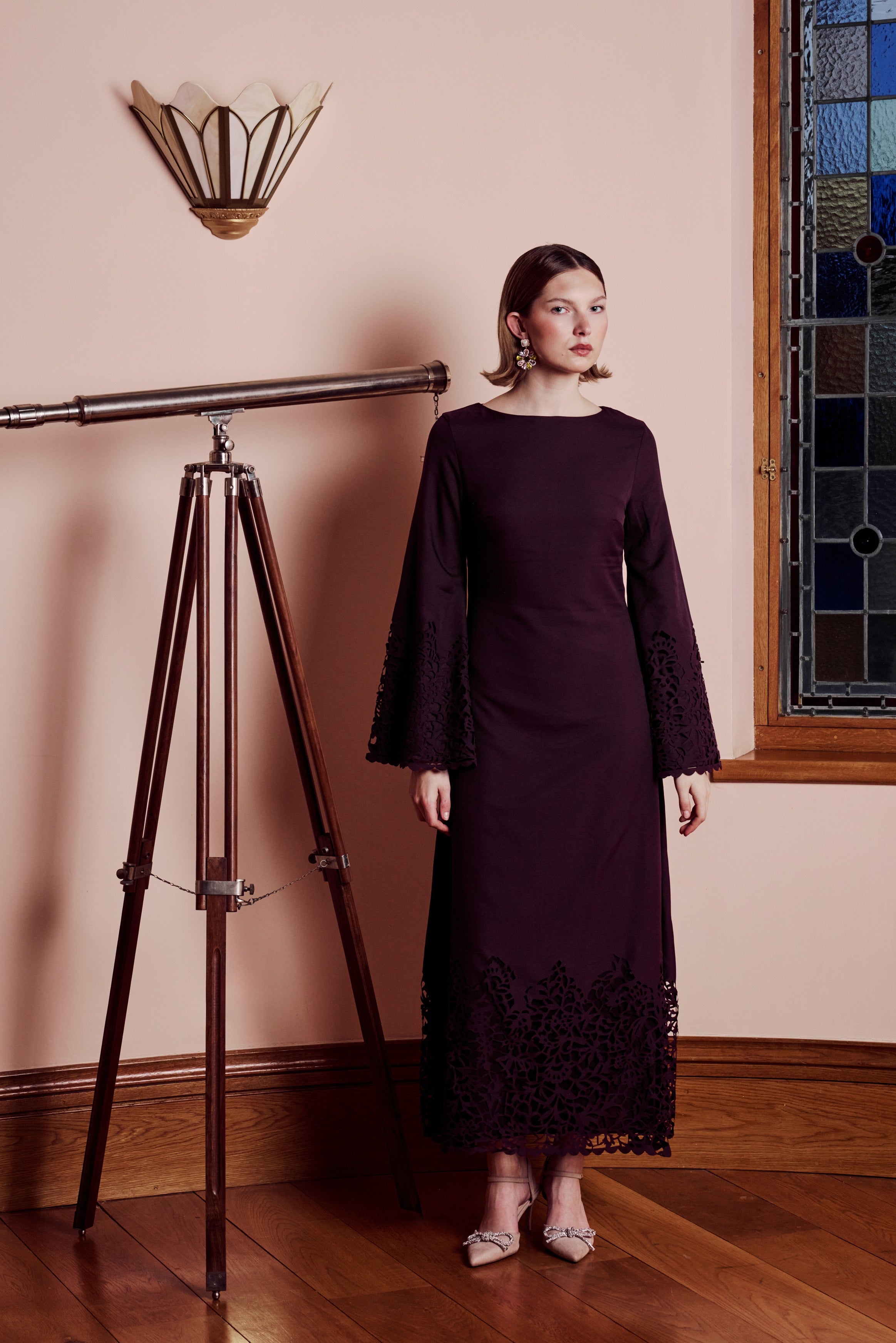 Woman in a long dark dress standing next to a telescope in a room with a stained glass window.
