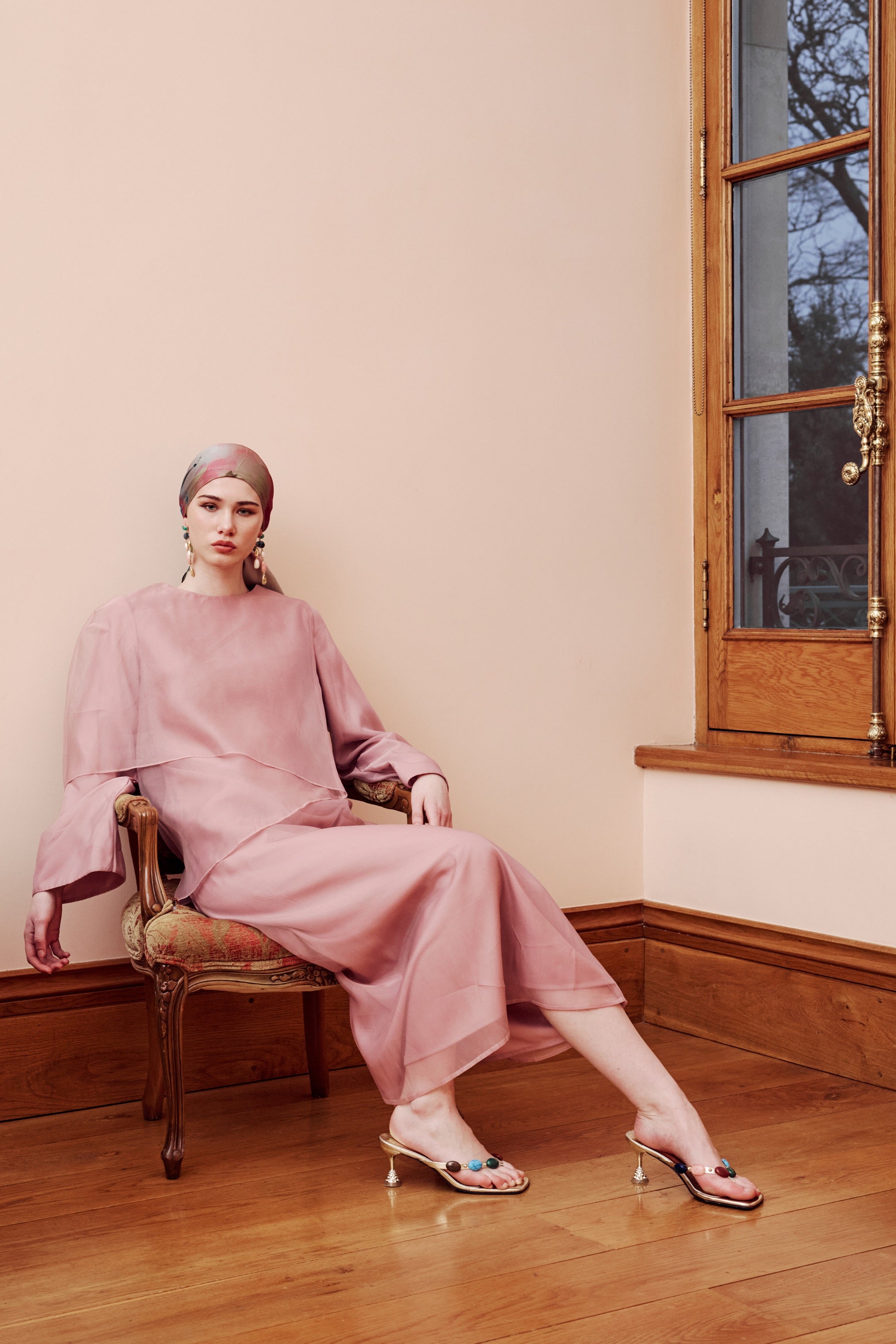 Woman in a pink outfit sitting on a chair in a room with wooden floor and window.