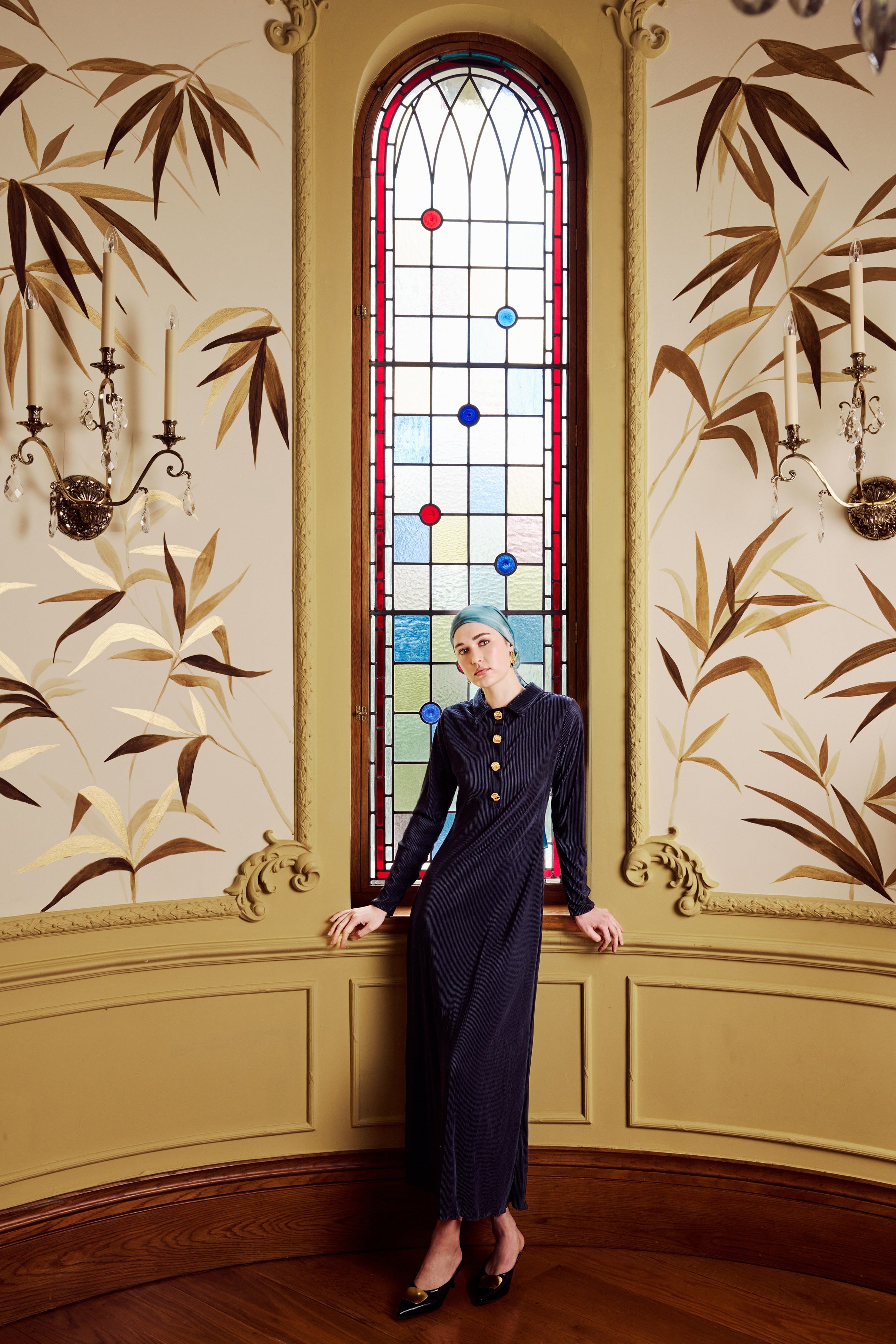Woman in a dark blue dress standing in front of a stained glass window with decorative wall panels.