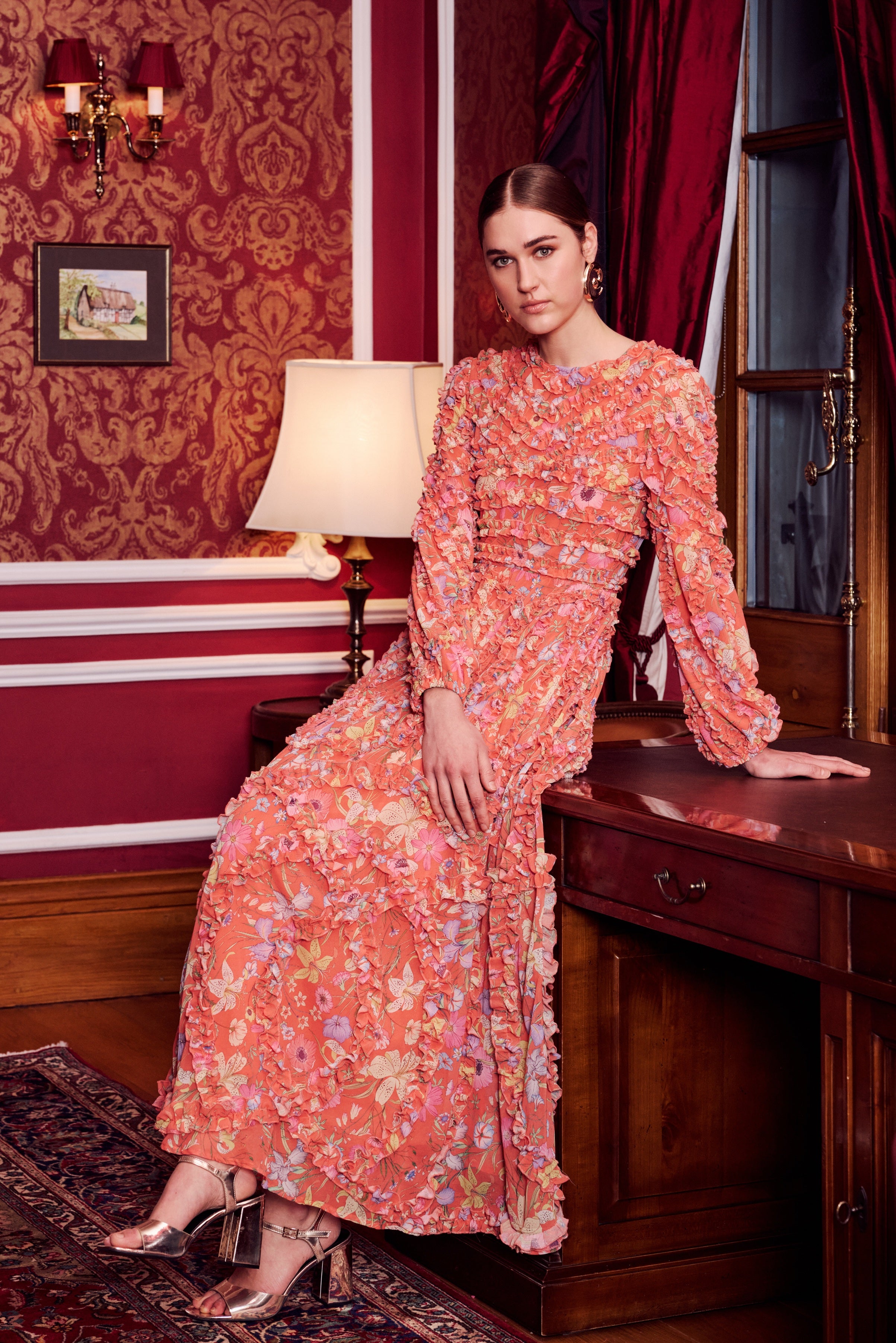 Woman in a floral dress standing in an elegant room with red walls and wooden furniture.
