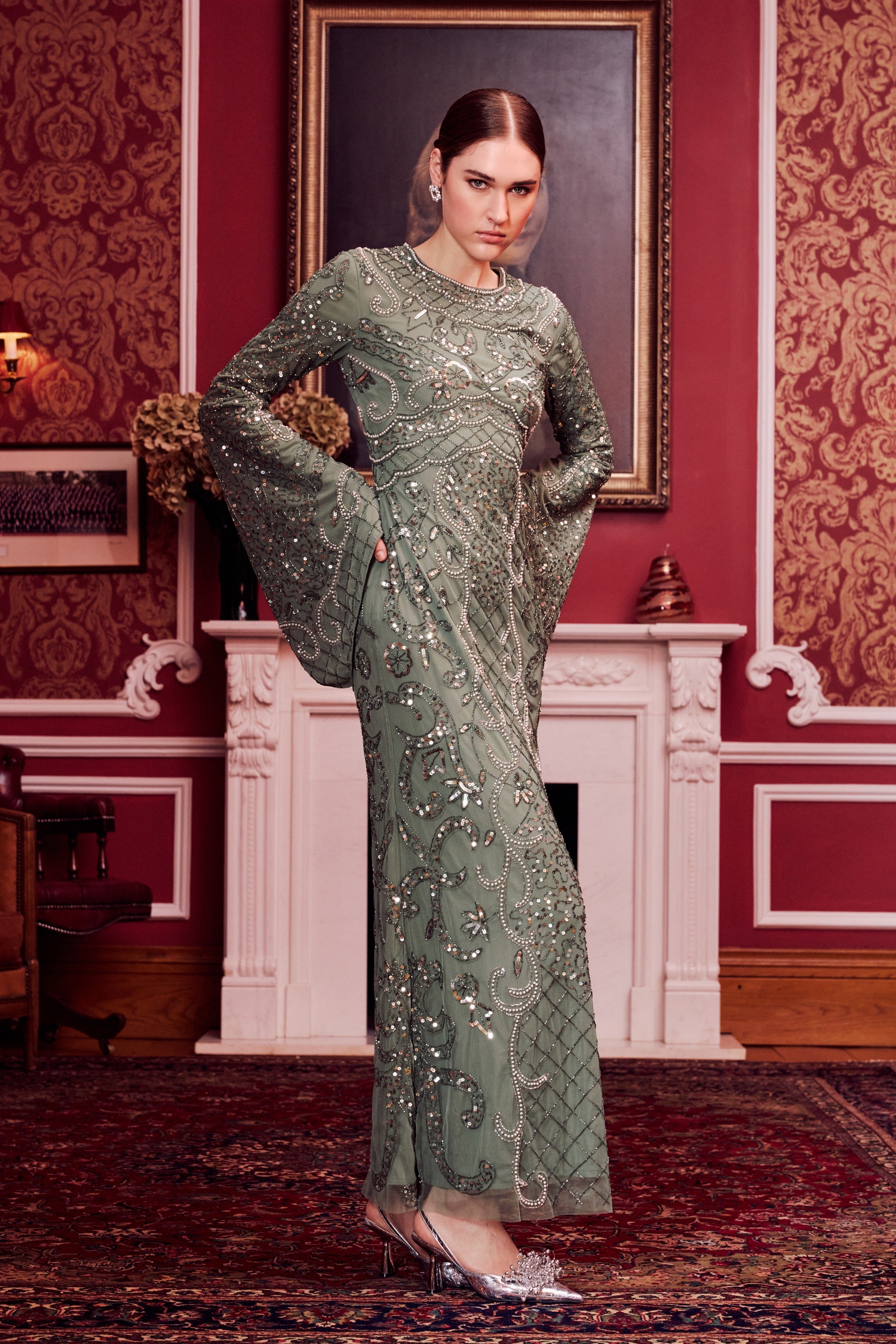 Woman in a green evening gown standing in a room with red wallpaper and a fireplace.