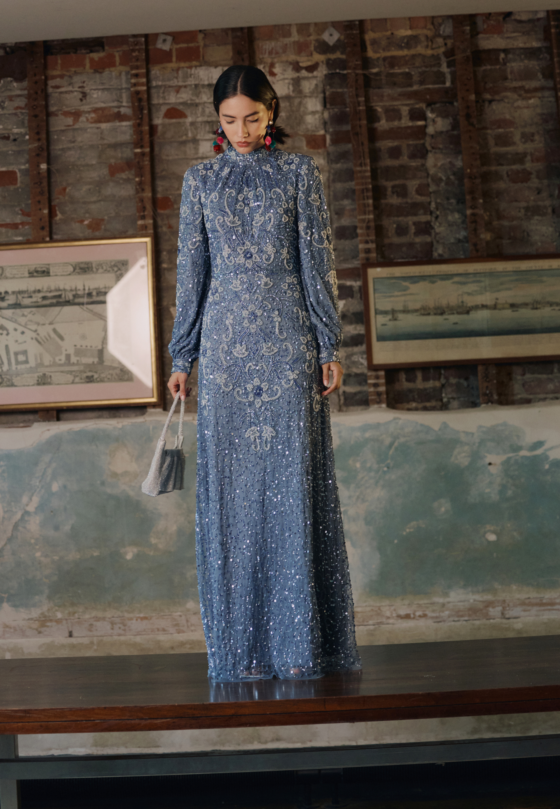 Woman in a blue lace dress standing in front of a textured wall with framed pictures.