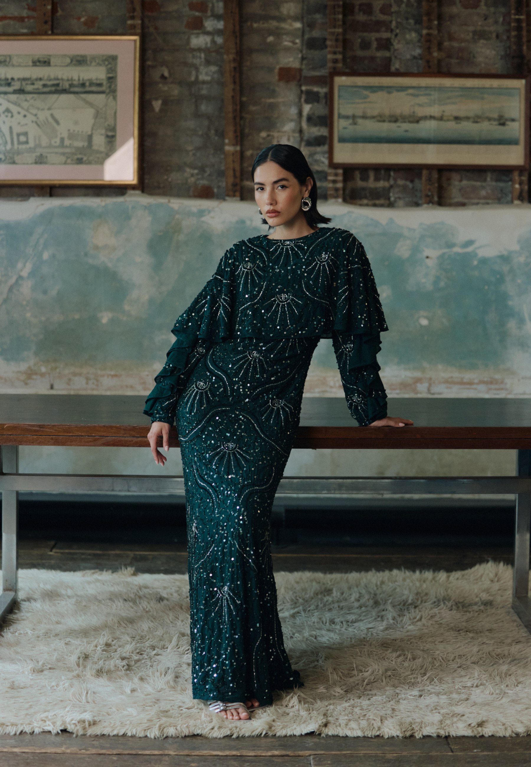 Woman in a dark green beaded dress standing in a room with a textured wall and bench.