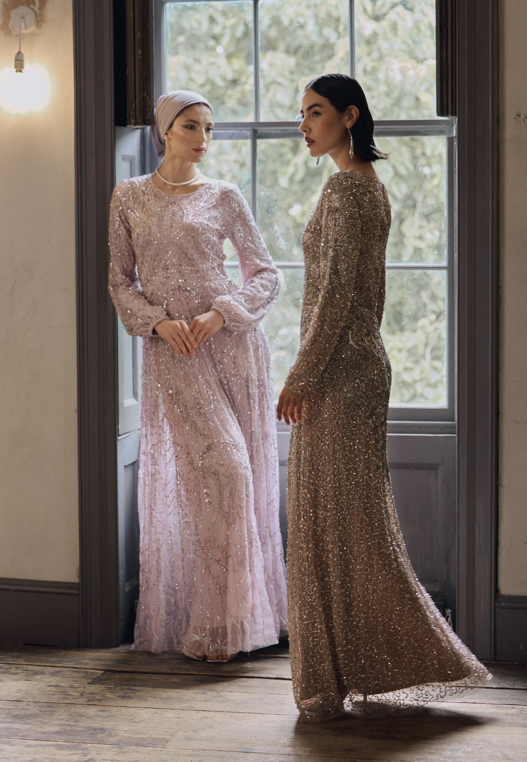 Two women in elegant evening gowns standing in a room with large windows.